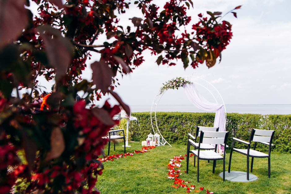 An outdoor wedding ceremony setup featuring a white circular floral arch decorated with green foliage and white flowers, positioned on a grassy area near a hedgerow with a distant view of the sea. In 