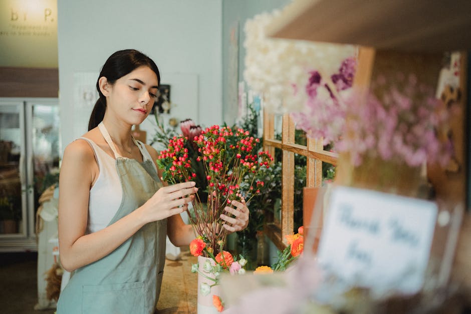 A young woman with fair skin and dark hair tied back, wearing a sleeveless white top and a beige apron, inspects a bouquet of pink and red waxflower stems in a floral shop. The bouquet features numero